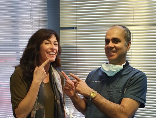 A smiling patient pointing to her teeth while standing next to a dentist in blue scrubs and a mask. The dentist is also pointing at her smile, both standing in front of window blinds in a dental clinic.