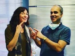 A smiling patient pointing to her teeth while standing next to a dentist in blue scrubs and a mask. The dentist is also pointing at her smile, both standing in front of window blinds in a dental clinic.
