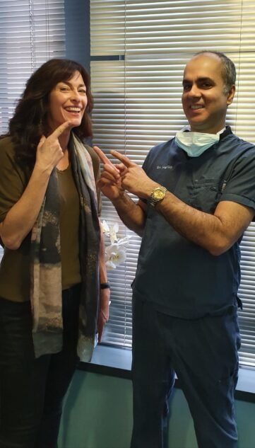 A smiling patient pointing to her teeth while standing next to a dentist in blue scrubs and a mask. The dentist is also pointing at her smile, both standing in front of window blinds in a dental clinic.