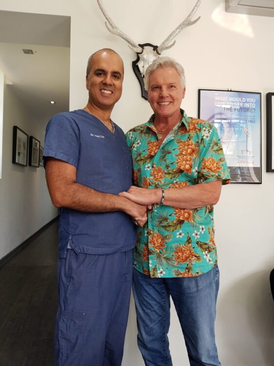 Our dentist and a patient holding hands, both smiling after a successful dental implants treatment. The dentist is wearing a blue coat while the patient has blue jeans and a green shirt with orange and white flowers.