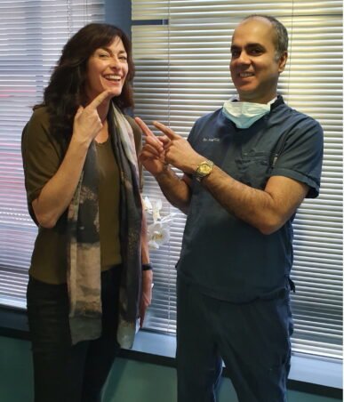 A patient and a dentist celebrating dental work results. The patient points to her teeth, and the dentist, in blue scrubs, points to her smile. Window blinds and a flower are visible in the background.