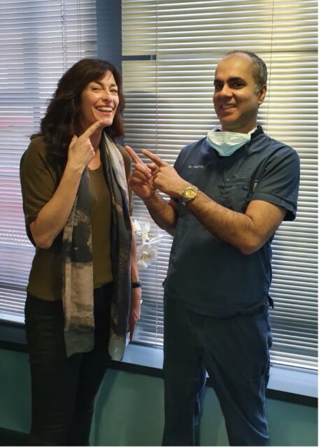 A patient and a dentist celebrating dental work results. The patient points to her teeth, and the dentist, in blue scrubs, points to her smile. Window blinds and a flower are visible in the background.
