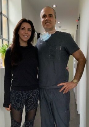 A female patient and our dentist posing together in our dental clinic hallway, both smiling at the camera. The dentist is wearing dark scrubs and a mask around his neck.
