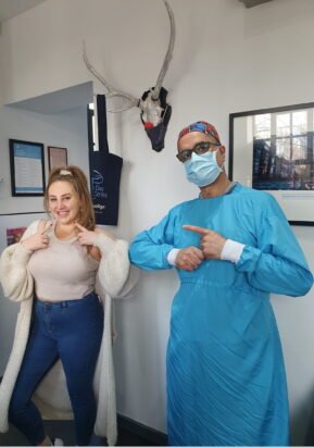 A smiling patient pointing to her teeth and our dentist in blue surgical scrubs and a mask pointing towards her, standing in our dental clinic with framed pictures and a decorative antler display on the wall.