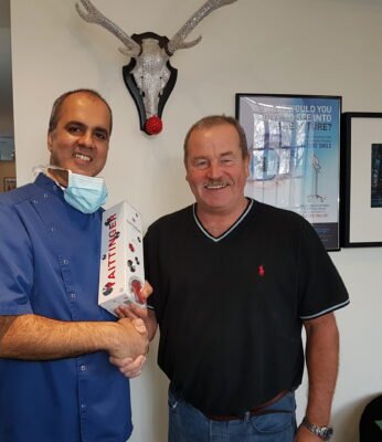 Out dentist and a patient posing together in our clinic, with the dentist wearing a blue coat and mask, while holding a thank-you gift after a successful dental implants treatment. The clinic wall features framed artwork and a decorative deer head.
